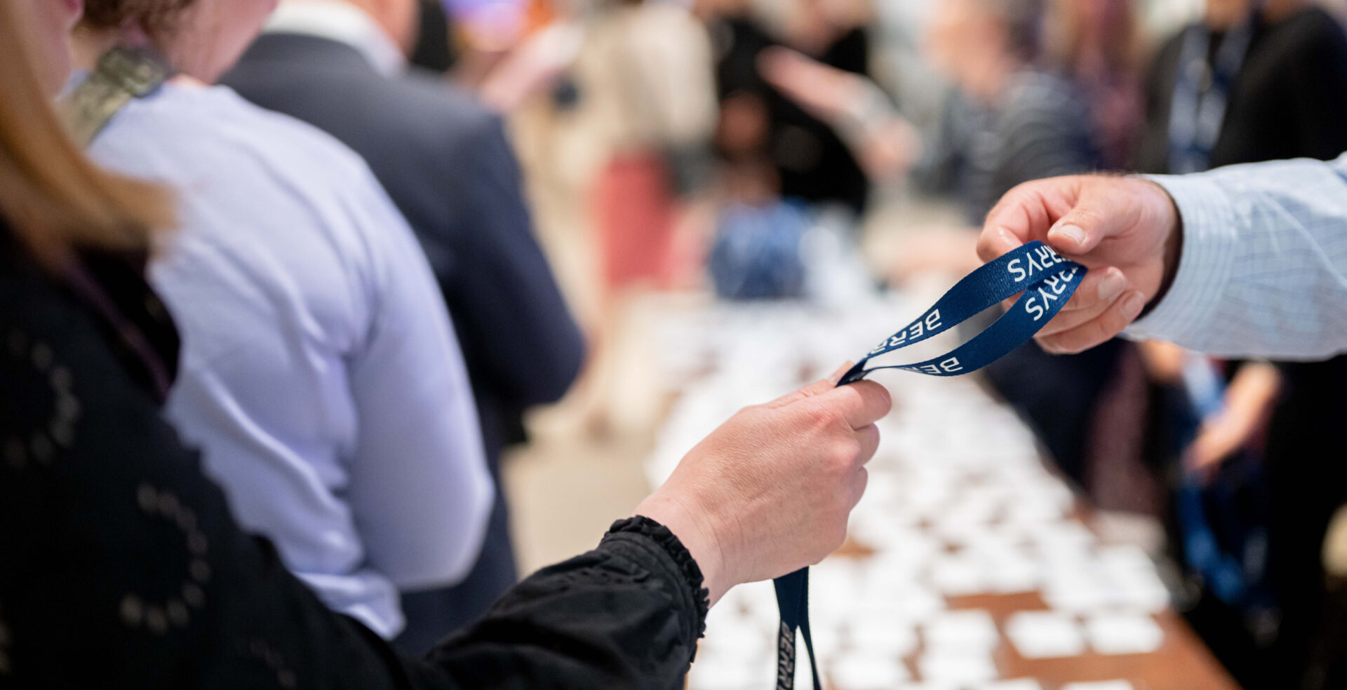 Close up of one person handing a blue event lanyard, that says Berrys to another, with a busy registration table and people in the background.