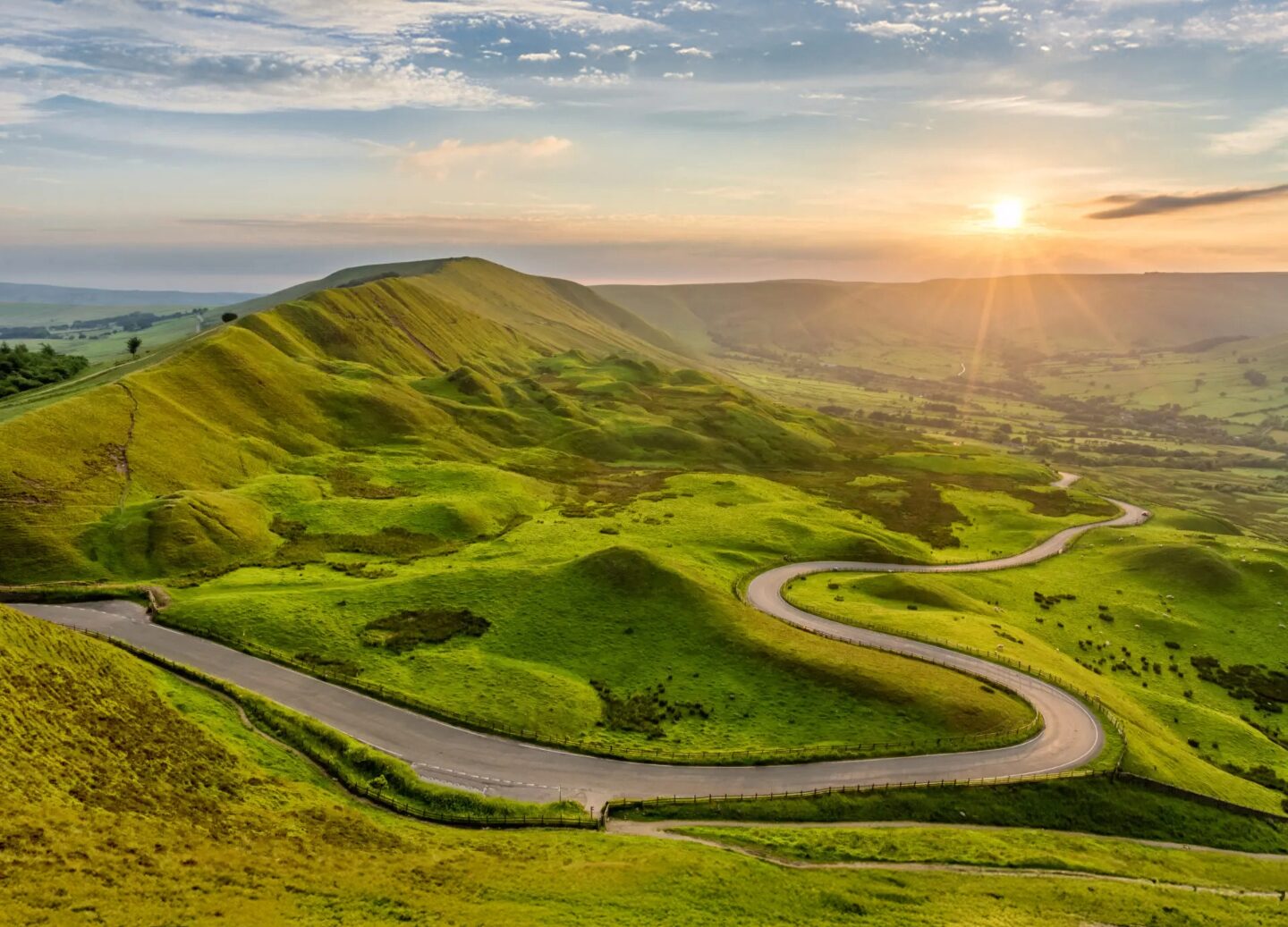 A curving country road weaves through vivid green rolling hills, with the sun rising over the horizon and casting a warm golden light across the landscape.