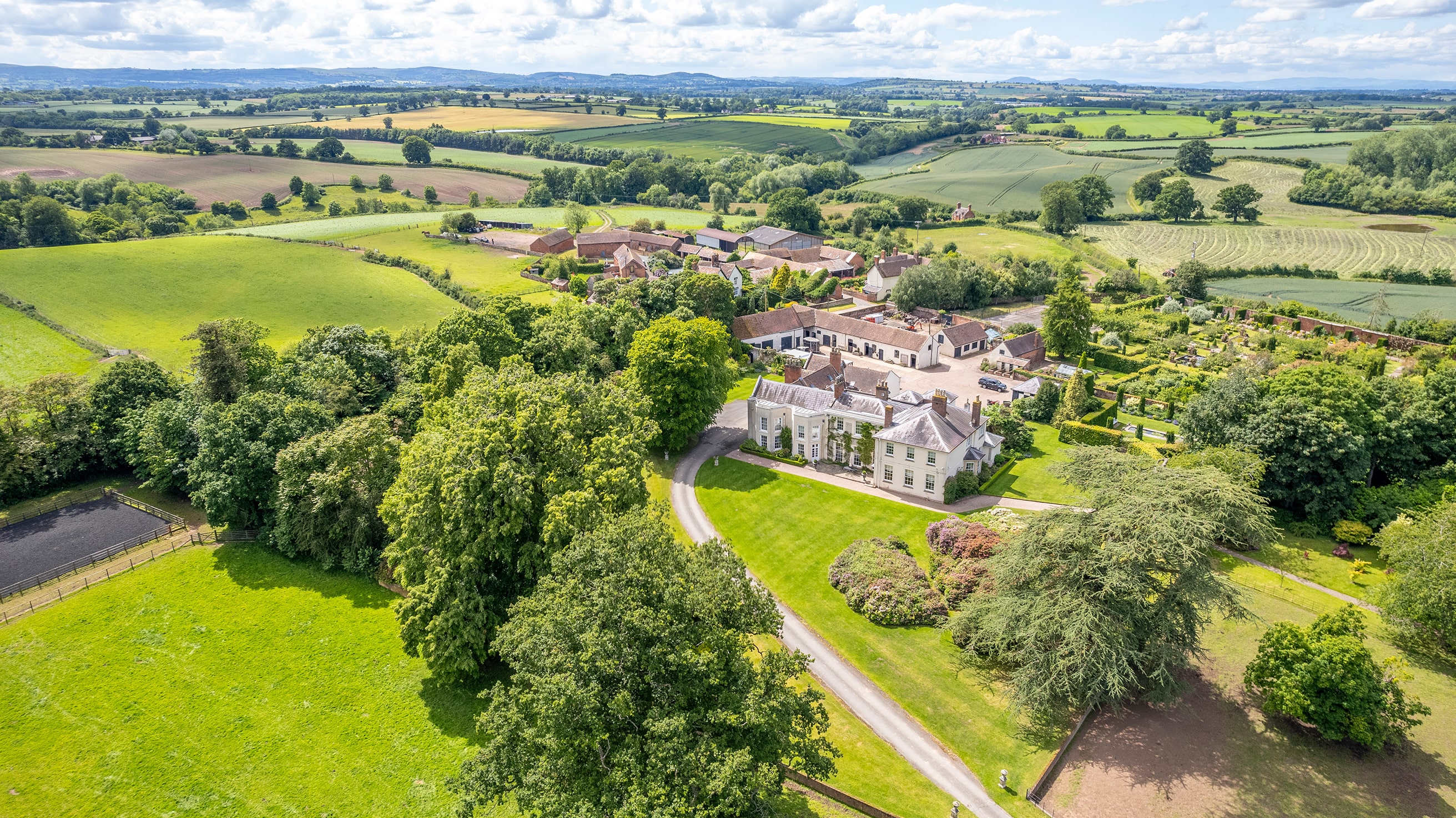 Elegant detached property with cream‑rendered walls, tall chimney stacks and dark grey slate roof under a bright blue sky. Nestled on a spacious drive, the home’s classic British architecture and tidy front lawn emphasise its premium residential appeal.